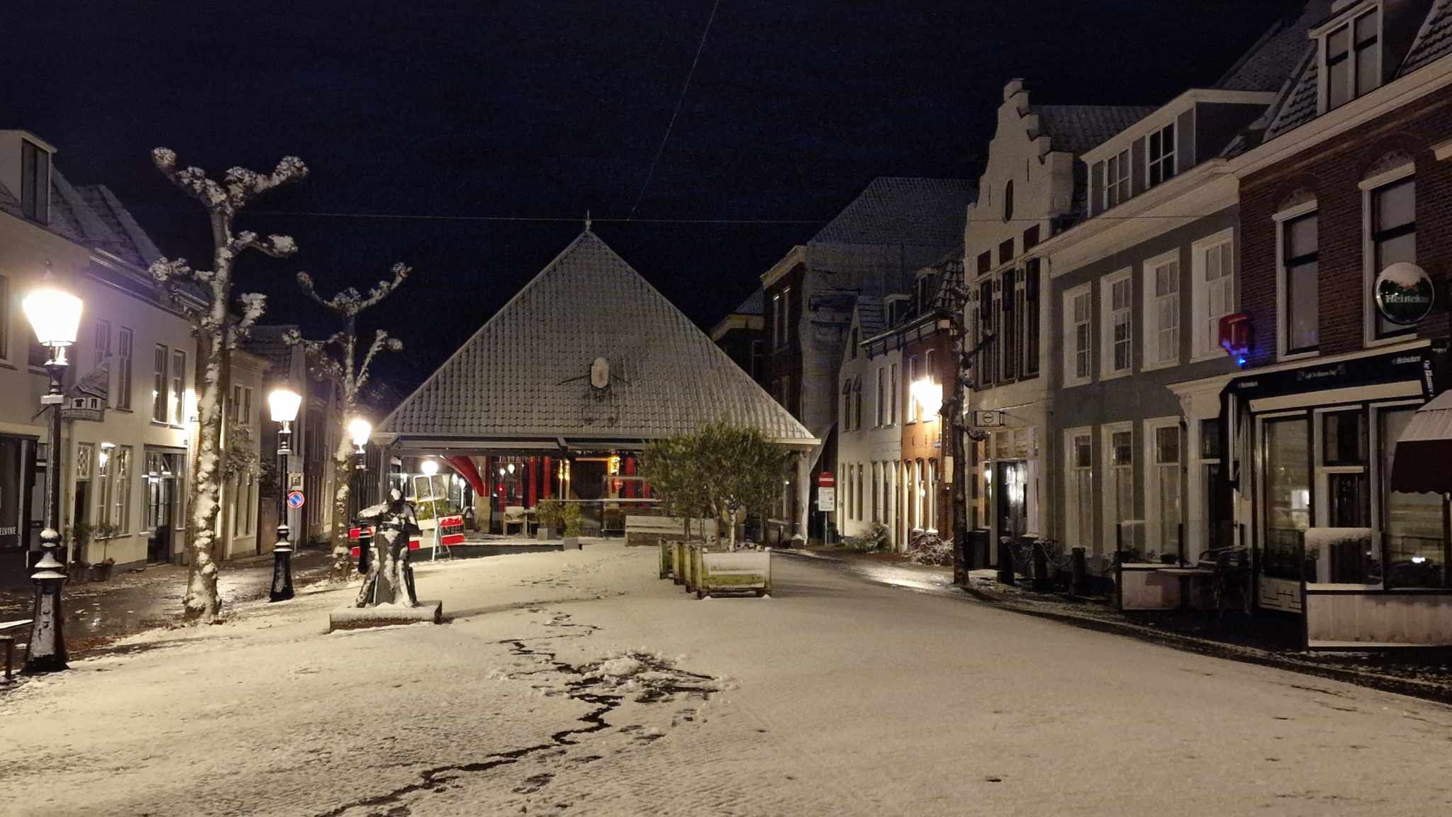 Een besneeuwd stadsplein in de avond, met brandende lantaarns en sneeuw op de bomen